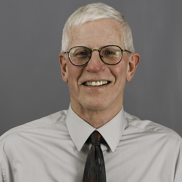 Larry Baker, An older man with short white hair, wearing glasses, a light gray shirt, and a dark tie, smiles against a plain gray background.