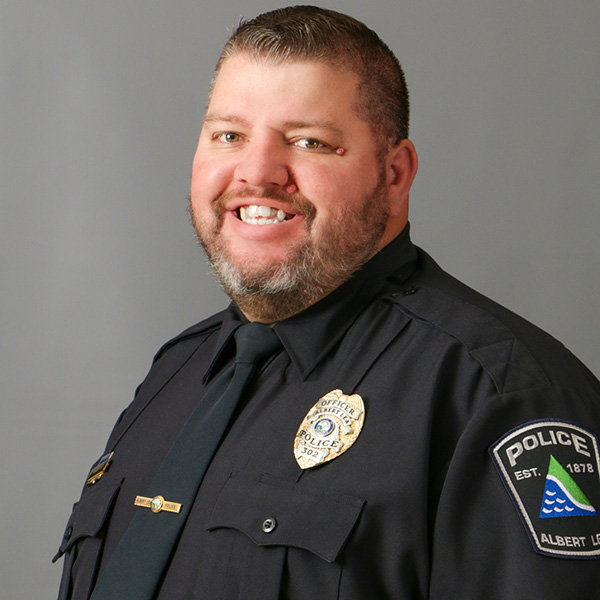 Luke Tuzinkski, Smiling man in a police uniform, featuring a badge and patches, posing against a neutral background.