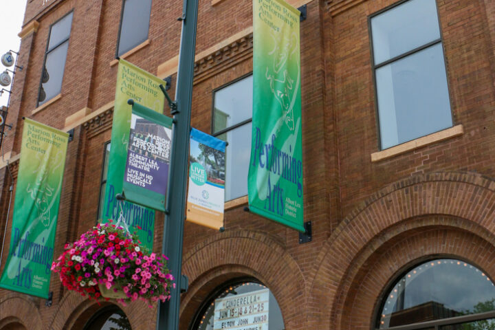Brick facade of the Marion Ross Performing Arts Center with banners, a streetlamp, hanging flower basket, and a bronze statue in the foreground.