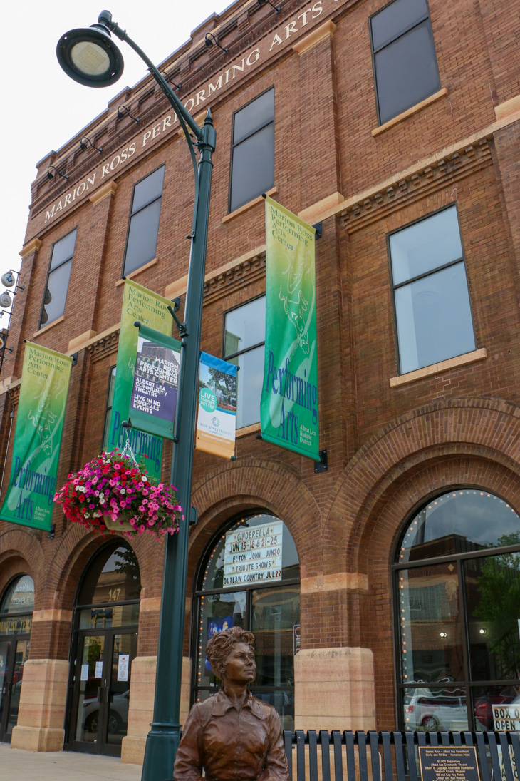 Brick facade of the Marion Ross Performing Arts Center with banners, a streetlamp, hanging flower basket, and a bronze statue in the foreground.