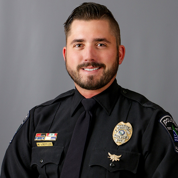 Matt Loeffler, A man in a police uniform smiles against a gray background. He wears badges on his shirt and has a dark tie.