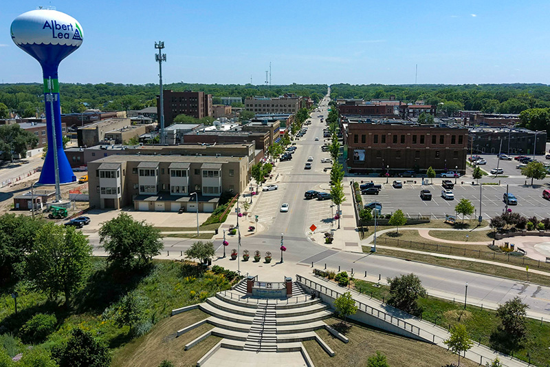 Aerial view of a small city with a water tower labeled "Albert Lea" and a main road lined with buildings, parking lots, and greenery on a clear day.