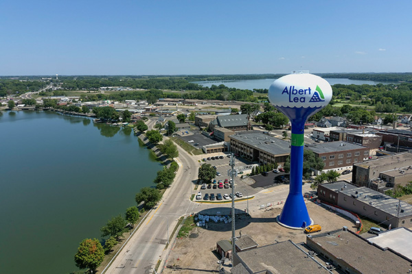A large water tower labeled "Albert Lea" stands near a lake, with buildings, roads, and cars surrounding it under a clear blue sky.