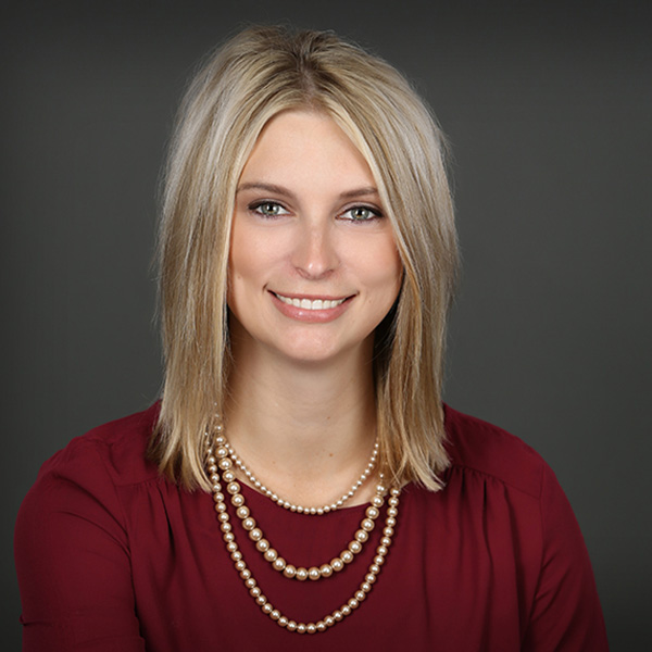 Megan Boeck, a Woman with straight blonde hair wearing a burgundy blouse and layered pearl necklaces, smiling at the camera against a plain dark background.