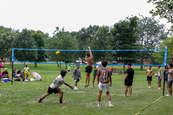 A group of people play volleyball on a grassy field while others sit and watch or relax in the background.