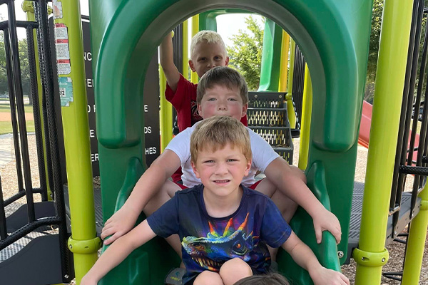 Four young boys sit in a row on a green playground slide, with climbing equipment and trees visible in the background.