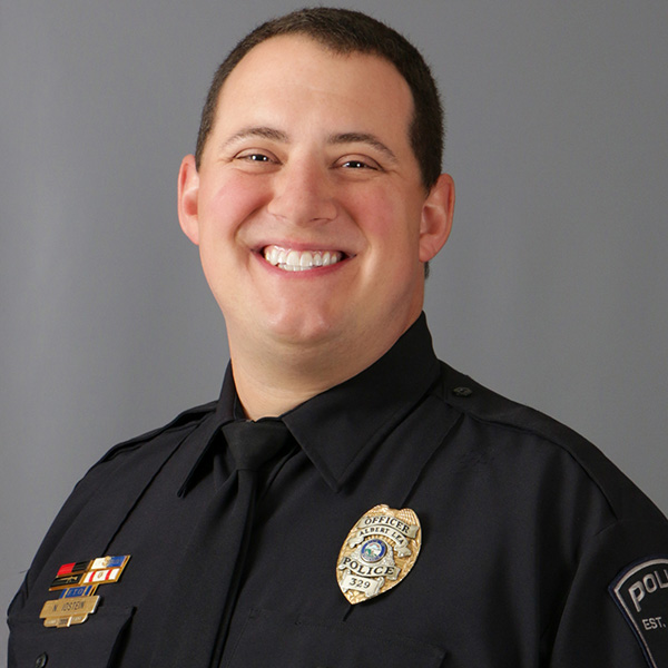 Nate Idstein, Smiling individual in a police uniform with badges, standing against a gray background.