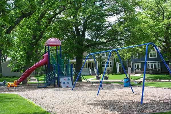 A playground with a slide, climbing wall, and swings is surrounded by trees and houses on a sunny day.