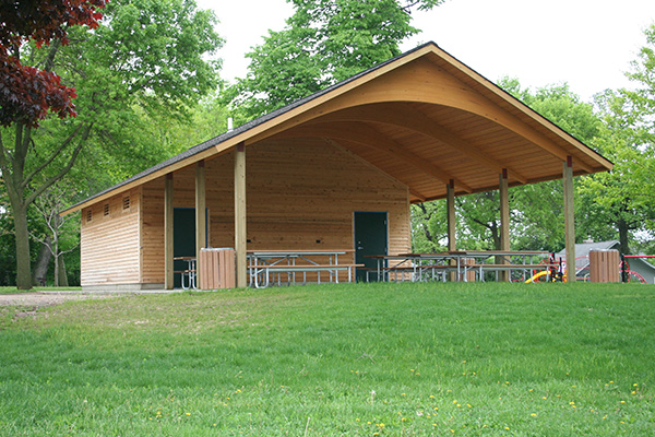 A wooden pavilion with a curved roof stands in a park, featuring picnic tables underneath and surrounded by green grass and trees.