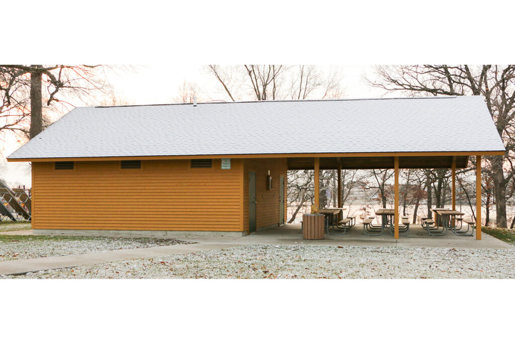 A wooden park shelter with a snow-covered roof, featuring an enclosed section and an open area with picnic tables underneath. Bare trees and a light dusting of snow are visible around the shelter.
