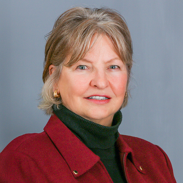 Rachel Christensen, Woman with short, light-colored hair, wearing a maroon jacket and a black turtleneck, against a gray background.