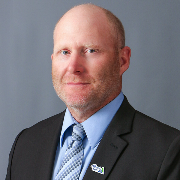 Reid Olson, A bald man with a beard, wearing a suit, light blue shirt, and patterned tie, is facing the camera against a gray background.