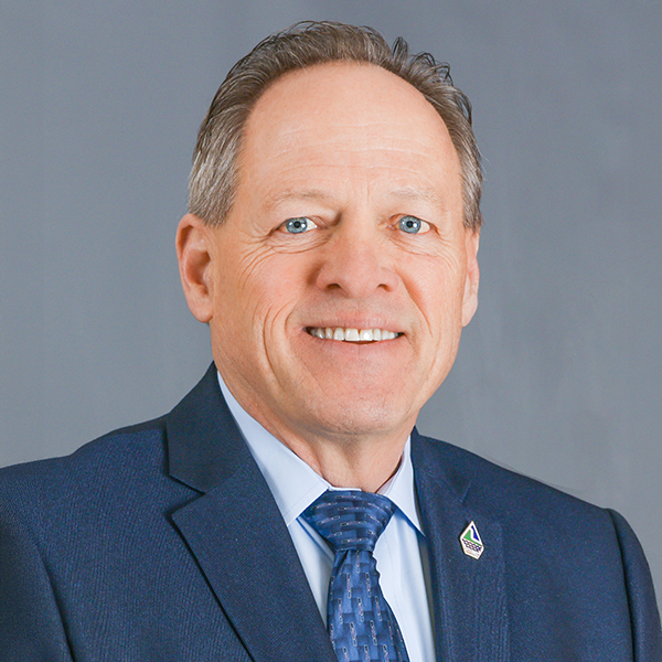 Rich Murray, Portrait of a man in a blue suit and tie, smiling, with a lapel pin on his jacket against a gray background.