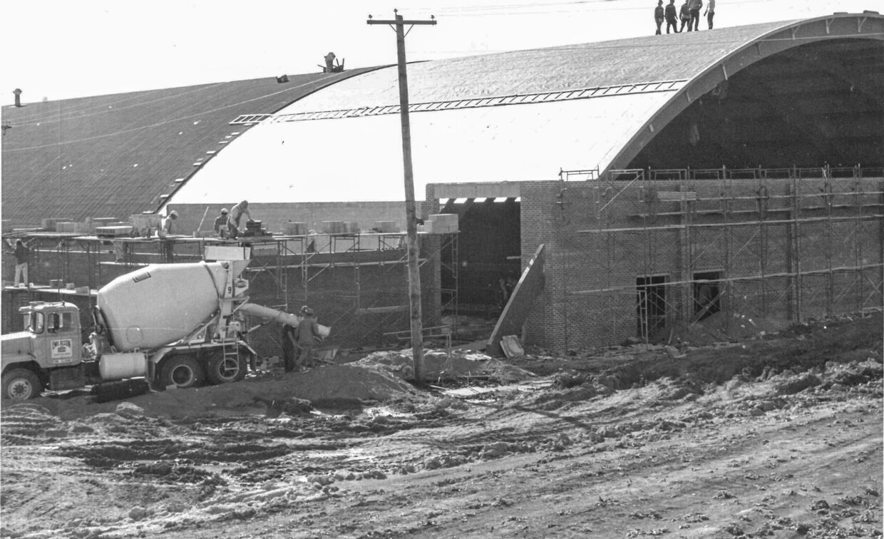 Construction site with workers on scaffolding and on the roof of a large arched building; cement truck parked in front and dirt ground in the foreground.