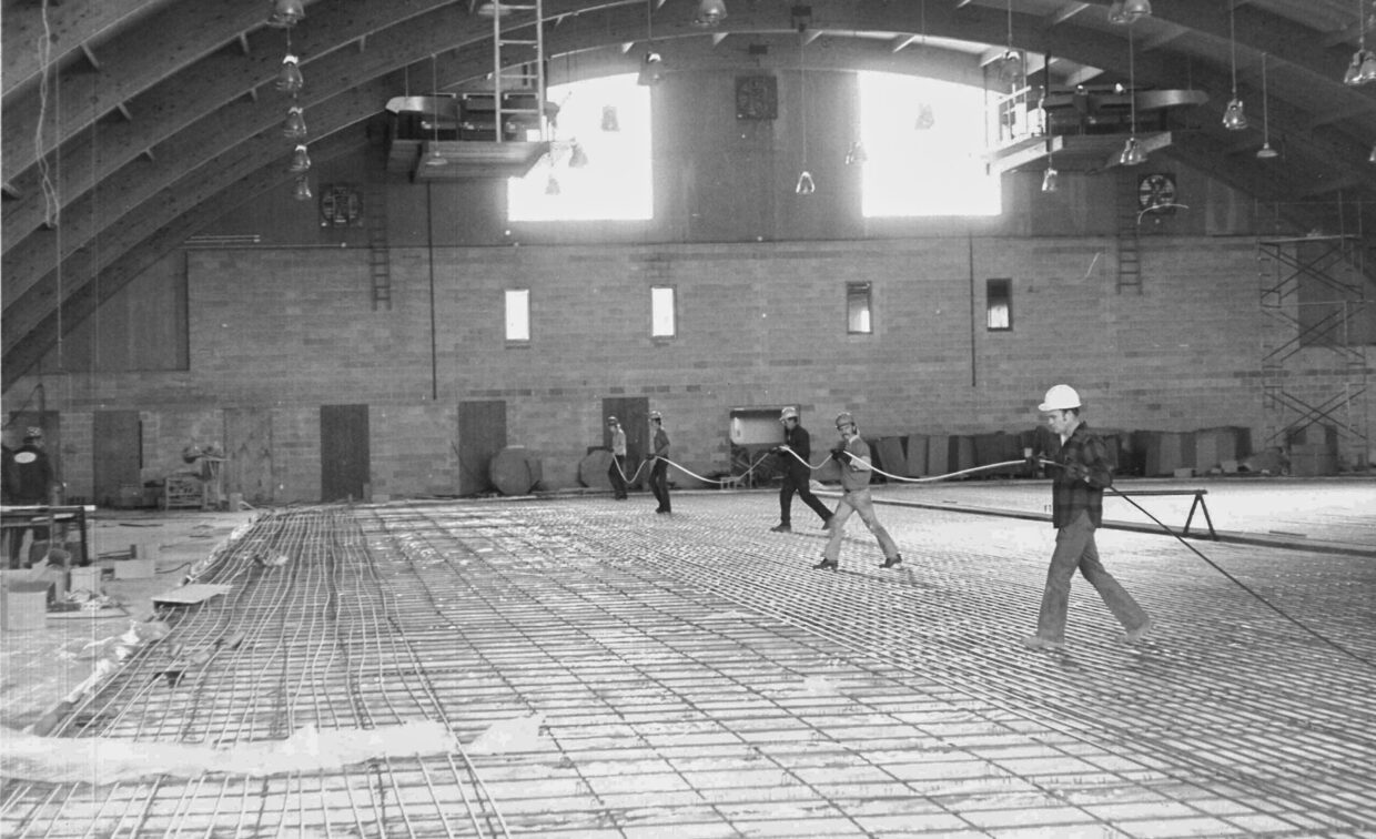 Construction workers installing rebar on the floor of a large indoor building with a curved wooden roof and exposed brick walls.