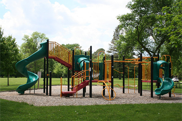 Playground with green and yellow slides, climbing structures, and ladders, set on wood chips in a grassy park with trees in the background.