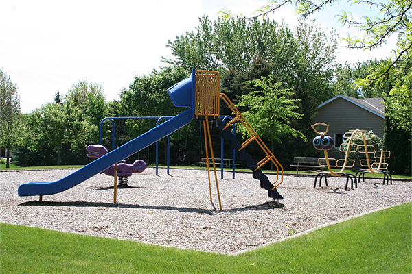 A playground with a blue slide, orange ladder, swings, spring riders, and climbing structures on a gravel surface, surrounded by trees and a house in the background.