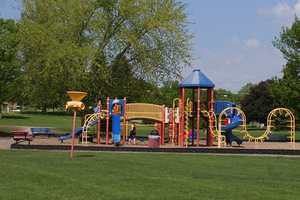 A colorful playground structure with slides and climbing equipment sits on a grassy field with trees and benches nearby under a partly cloudy sky.