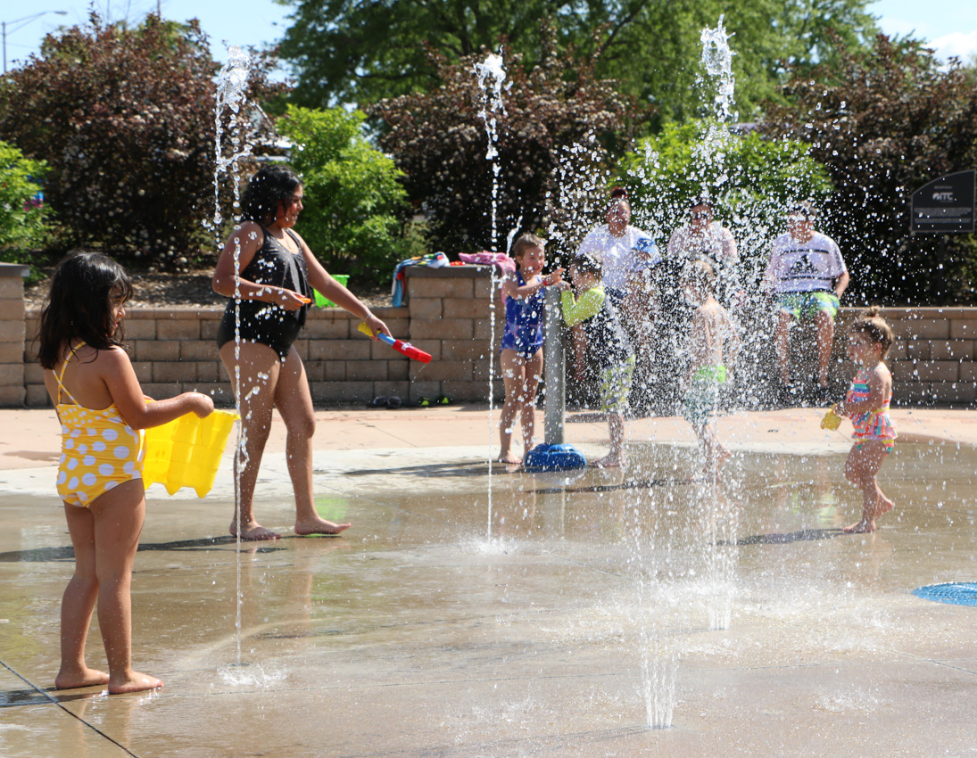 Children play in swimsuits among water jets at a splash pad, while several adults sit on a nearby wall in the background.