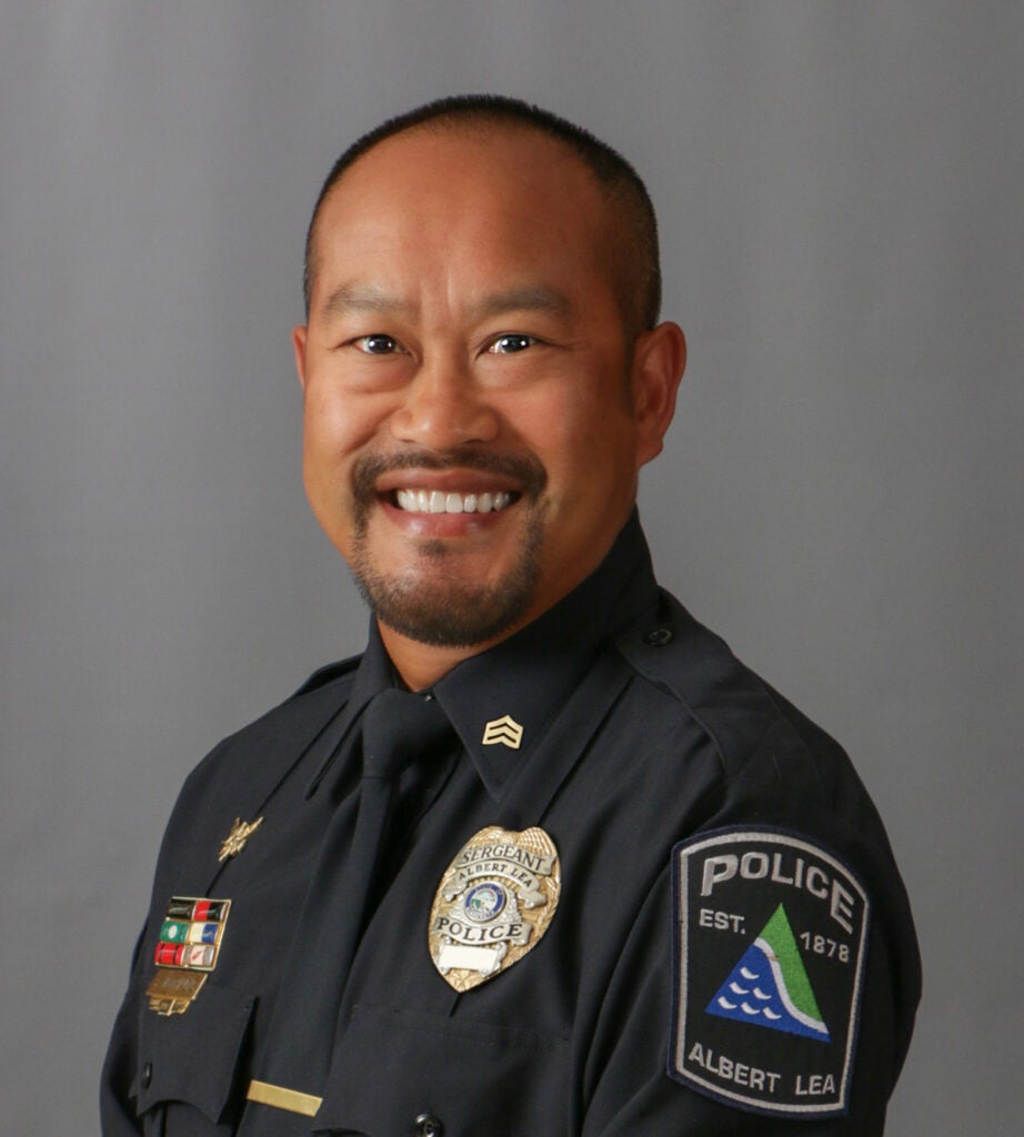 Sy Vanthavong, Smiling police officer in uniform with badges on the chest and arm, set against a gray background.