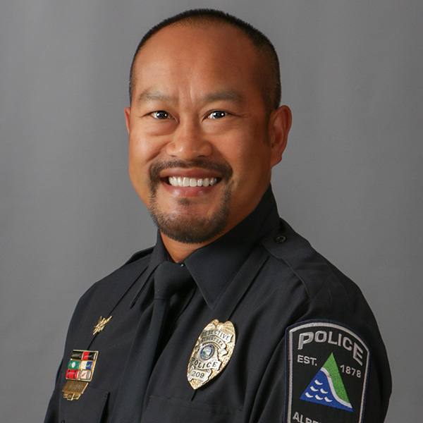 Sy Vanthavong, Smiling police officer in uniform with badges on the chest and arm, set against a gray background.