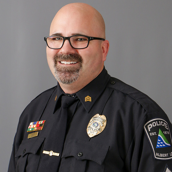 Tim Harves , A man wearing glasses and a police uniform, featuring badges and a sergeant insignia, smiles against a gray backdrop.