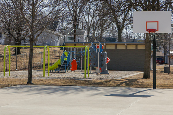 A playground with swings, a slide, climbing equipment, and a basketball hoop on a concrete court in a park with leafless trees.