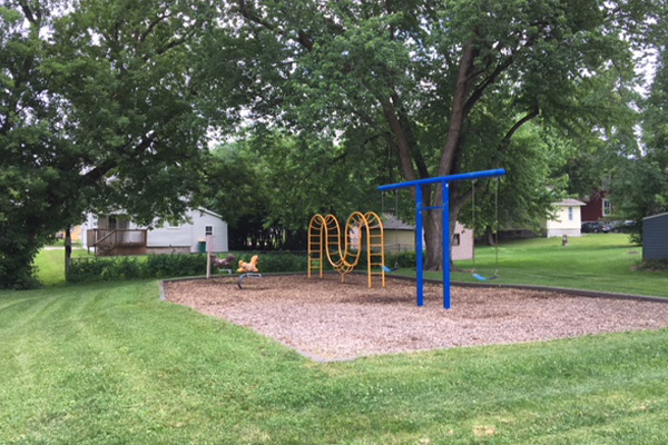 A playground with a blue swing set, yellow climbing bars, a spring rider, and wood chip ground, surrounded by grass and trees, with houses in the background.