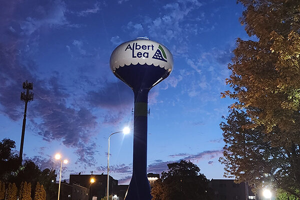 A water tower labeled "Albert Lea" stands against a twilight sky, surrounded by trees and city buildings, with streetlights illuminating the area.