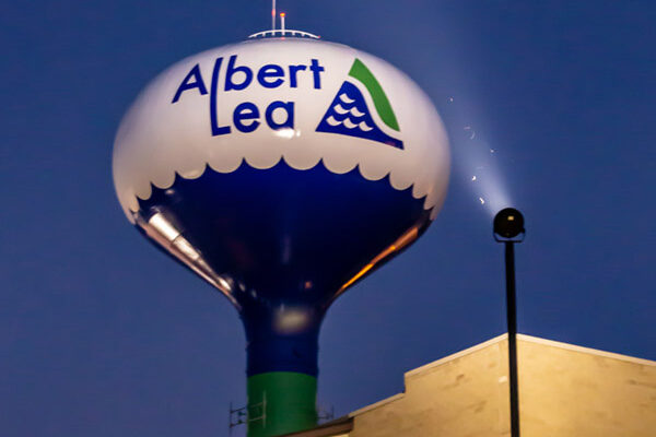 A large water tower at dusk displays the words "Albert Lea" with a green and blue wave logo, standing next to a beige building.
