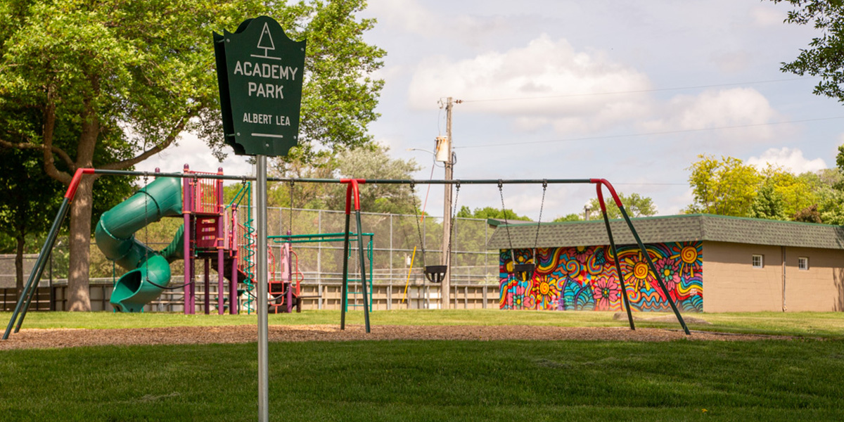 A playground at Academy Park with a slide, swings, grassy area, and a building featuring a colorful mural under a partly cloudy sky.
