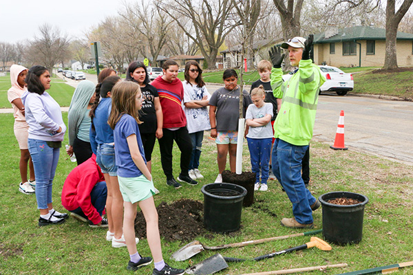 A group of children listens to an adult giving instructions near two planting holes and garden tools on a grassy area beside a street.