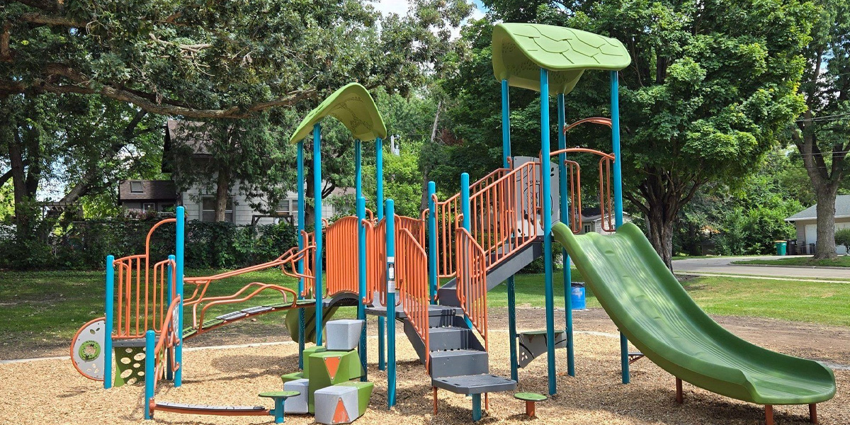 Outdoor playground set with slides, climbing steps, and green and orange features, surrounded by trees and houses, on a bed of wood chips.