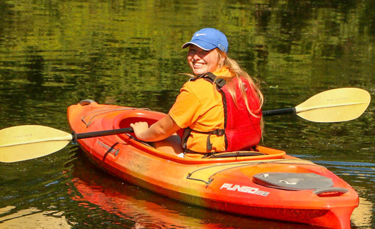 A person wearing an orange shirt, red life jacket, and blue cap paddles an orange kayak on calm water, looking back and smiling.