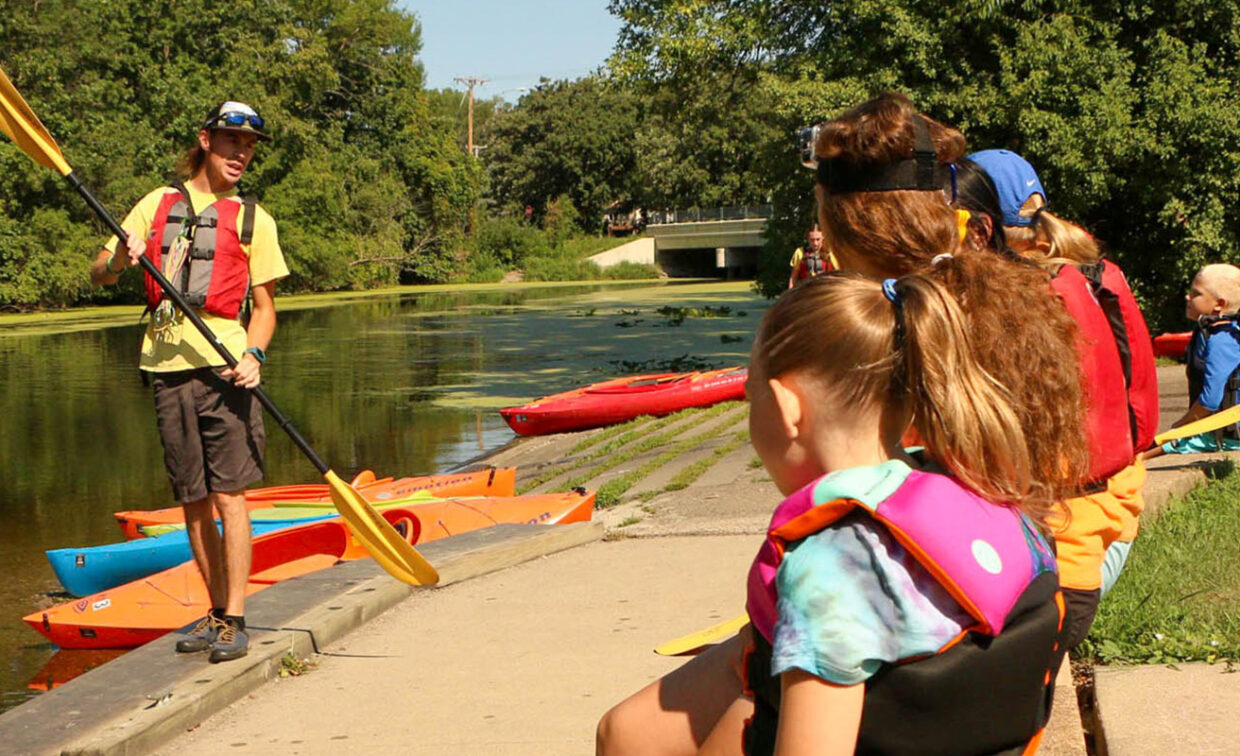 A group of children in life jackets listen to an adult instructor demonstrating how to hold a kayak paddle by the edge of a river, with kayaks lined up nearby.