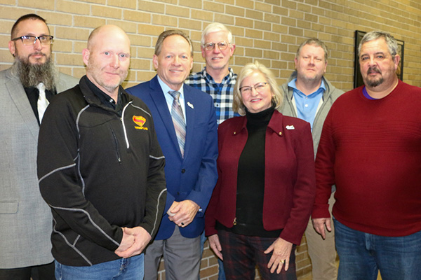 Seven adults, four men in casual or business attire and three in jackets or sweaters, stand in a row smiling in front of a tan brick wall.