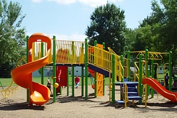 A colorful outdoor playground with slides, climbing bars, and ladders stands on a sandy surface surrounded by green trees.