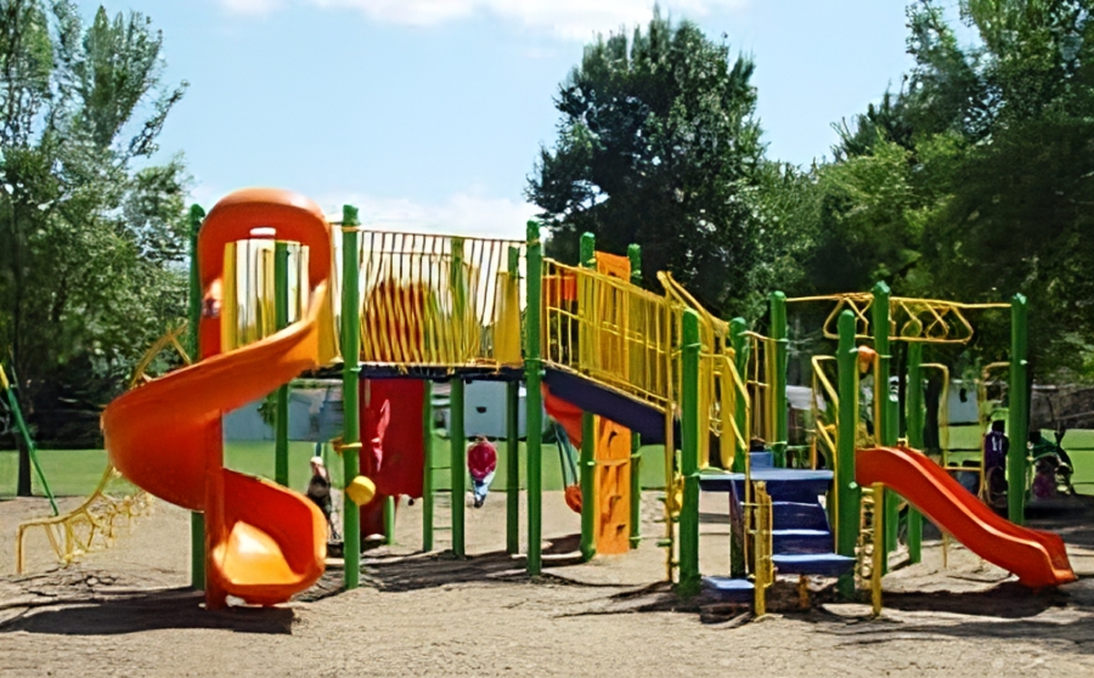 A colorful outdoor playground with slides, climbing bars, and ladders stands on a sandy surface surrounded by green trees.