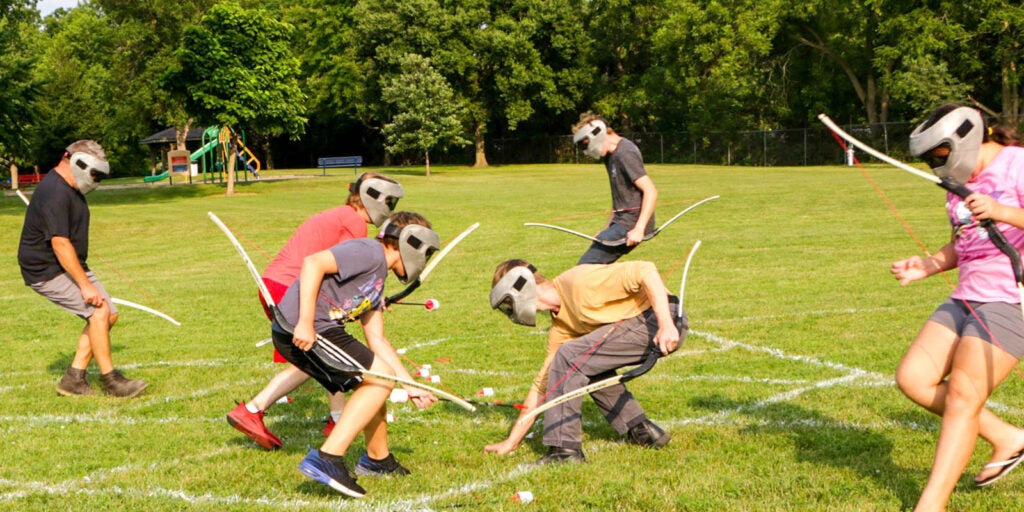 A group of children wearing protective masks play archery tag on a grassy field, holding bows and picking up foam-tipped arrows.