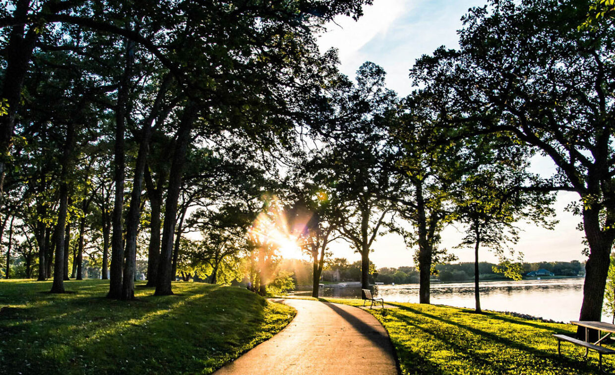 Paved path through a park with tall trees, grass, and a bench near a lake at sunset. Sunlight filters through the branches.