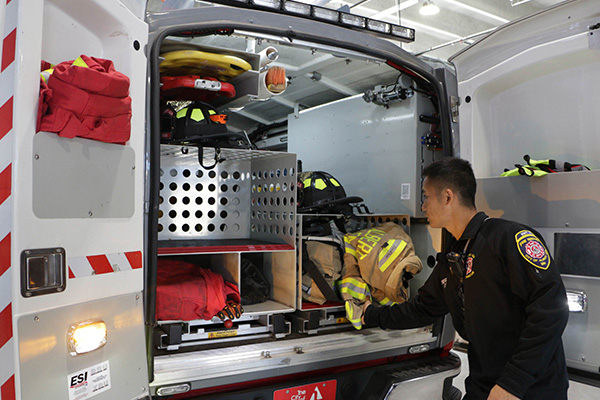 A firefighter organizes gear inside the open back compartment of an emergency response vehicle.
