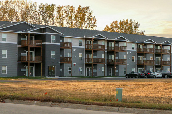 Three-story apartment building with balconies, gray siding, and parked cars in front; trees and a grassy area are visible in the background.