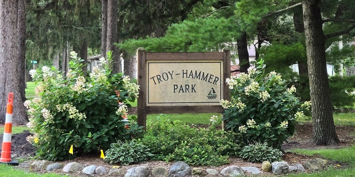 A wooden sign reading "Troy-Hammer Park" is surrounded by green bushes and trees; a traffic cone is visible on the left side.