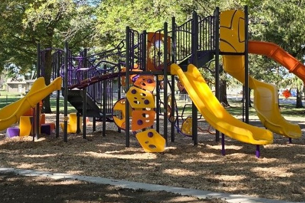 A playground structure with multiple yellow slides, climbing features, and platforms is set on wood chips and surrounded by trees in a park.