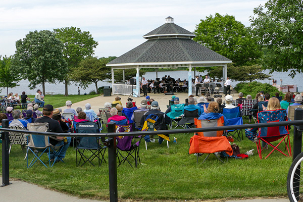 People sit in lawn chairs facing a band performing in a gazebo near a body of water, with trees and grass surrounding the area.