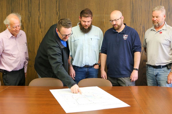 5 men look at item on tabletop, one man pointing to something