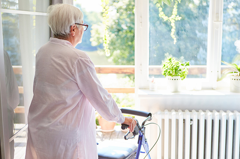 An elderly woman using a walker looks outside on a beautiful day.