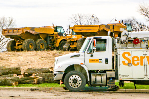 A white service truck in the foreground, with two large yellow dump trucks and a pile of logs in the background at a construction site.