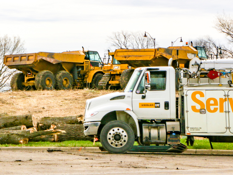 A white service truck in the foreground, with two large yellow dump trucks and a pile of logs in the background at a construction site.