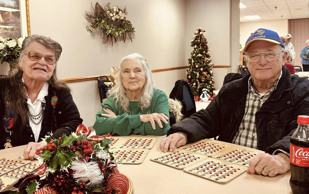 Query successful Three senior citizens are seated at a table, likely playing bingo, as bingo cards with red markers are visible in front of them. On the left, a woman with glasses and dark hair smiles at the camera. In the center, another woman with long grey hair and a green sweater looks towards the camera. On the right, a man wearing a blue "Vikings" baseball cap and a dark jacket also smiles at the camera.